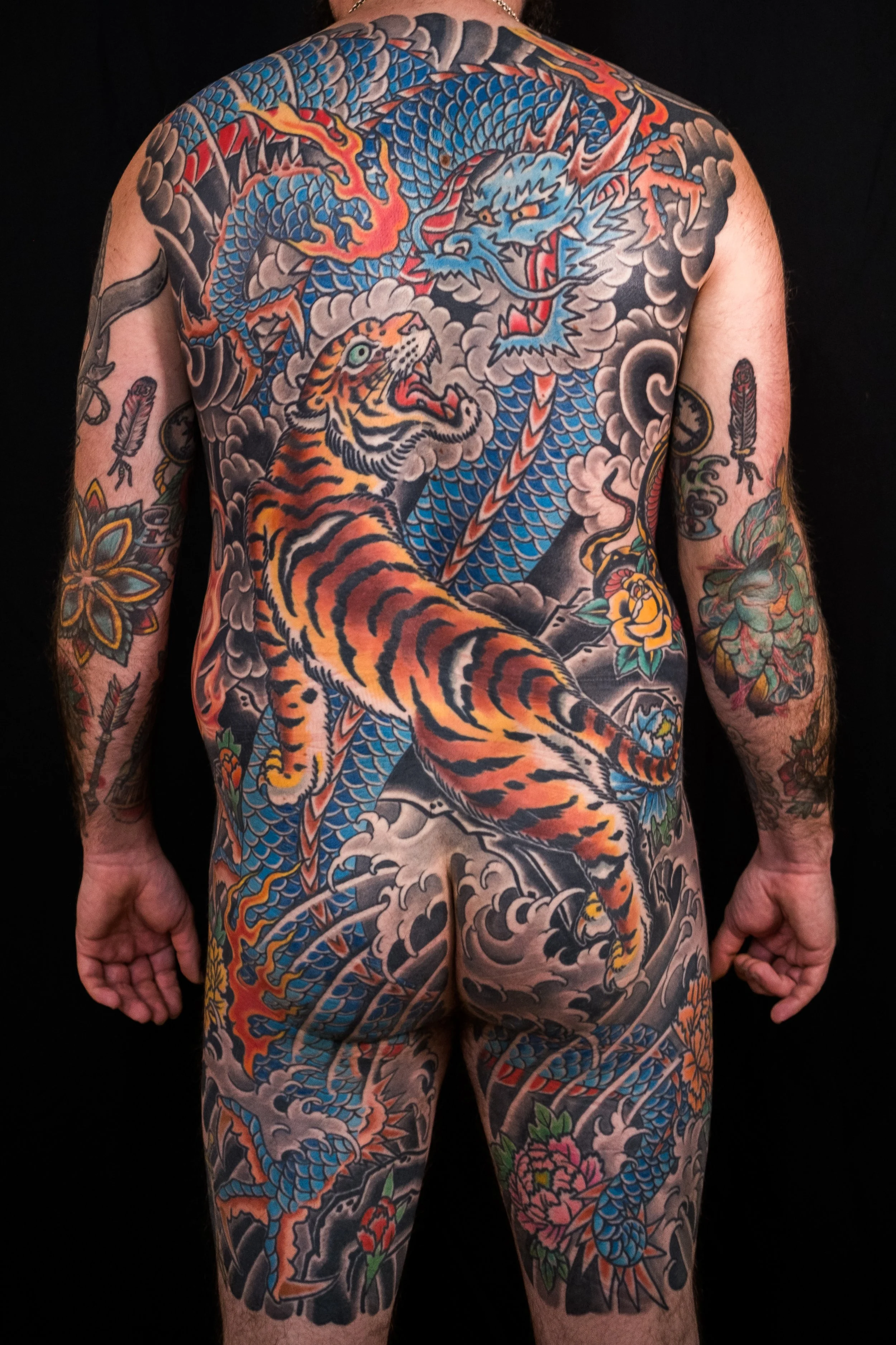 A person with a large full-back tattoo of a blue and orange dragon intertwined with a tiger, surrounded by black clouds and colorful flowers on arms.