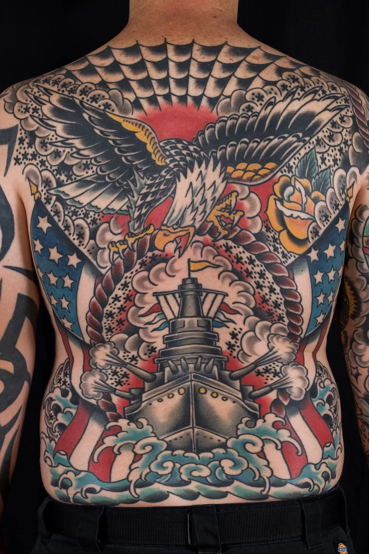Colorful tattoo of an eagle soaring above a battleship, with clouds, sky, and a red background.