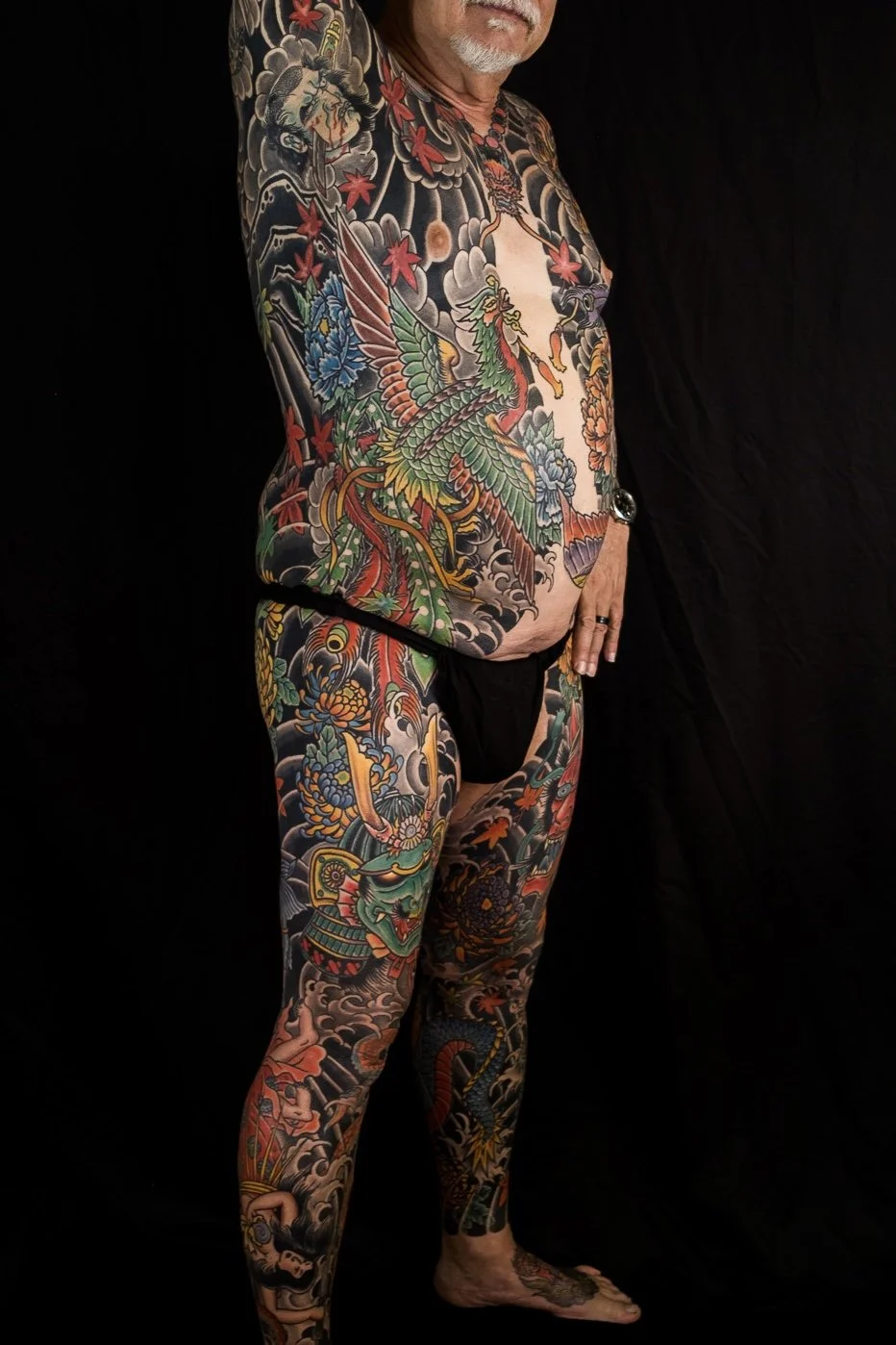 A man with extensive colorful tattoos covering his upper body, arms, torso, legs, and feet, standing against a black background. He has a gray beard and is wearing black underwear.