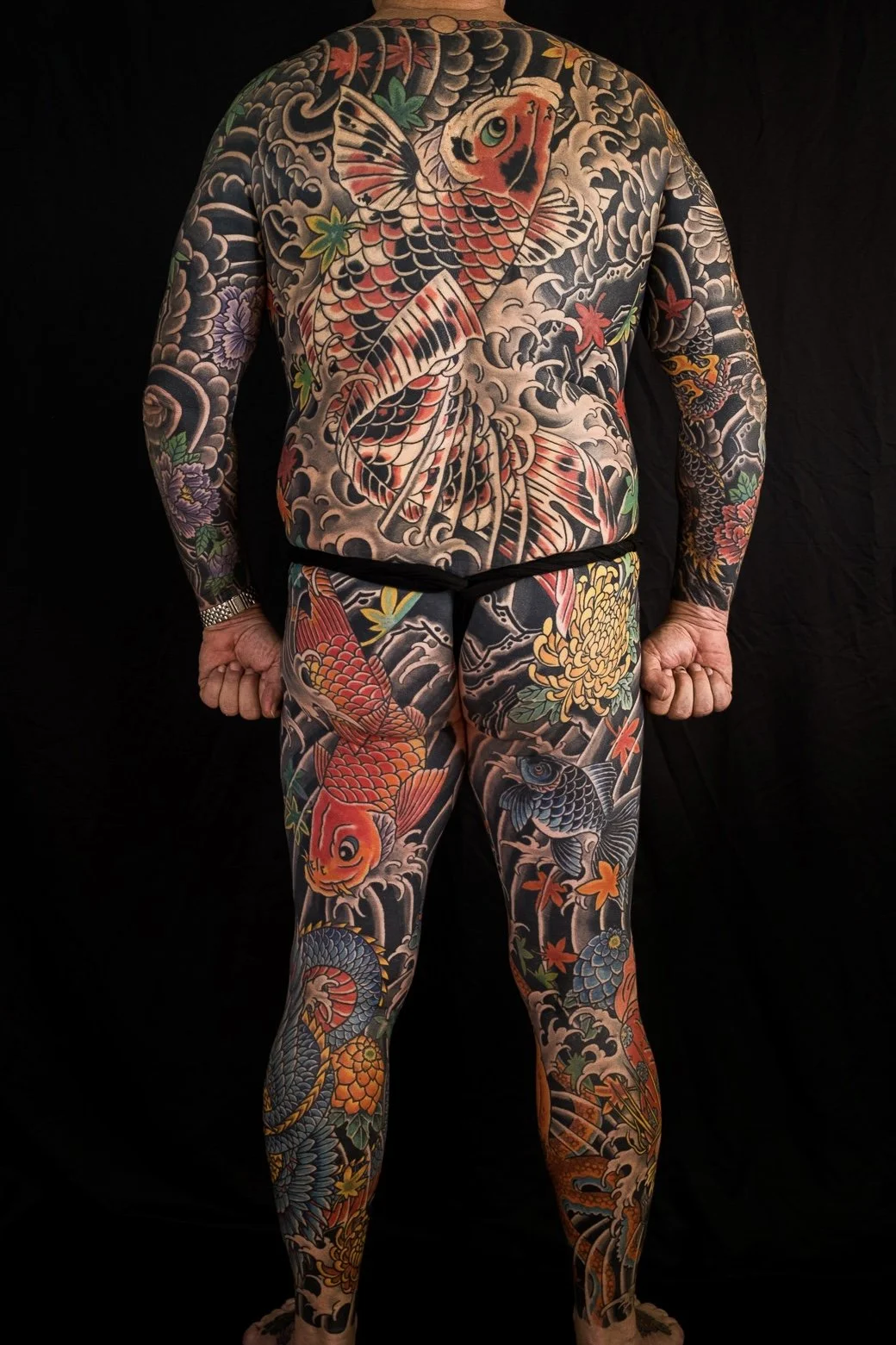 A person with a full-body Japanese-style tattoo featuring koi fish, waves, and colorful flowers, covering most of their skin.