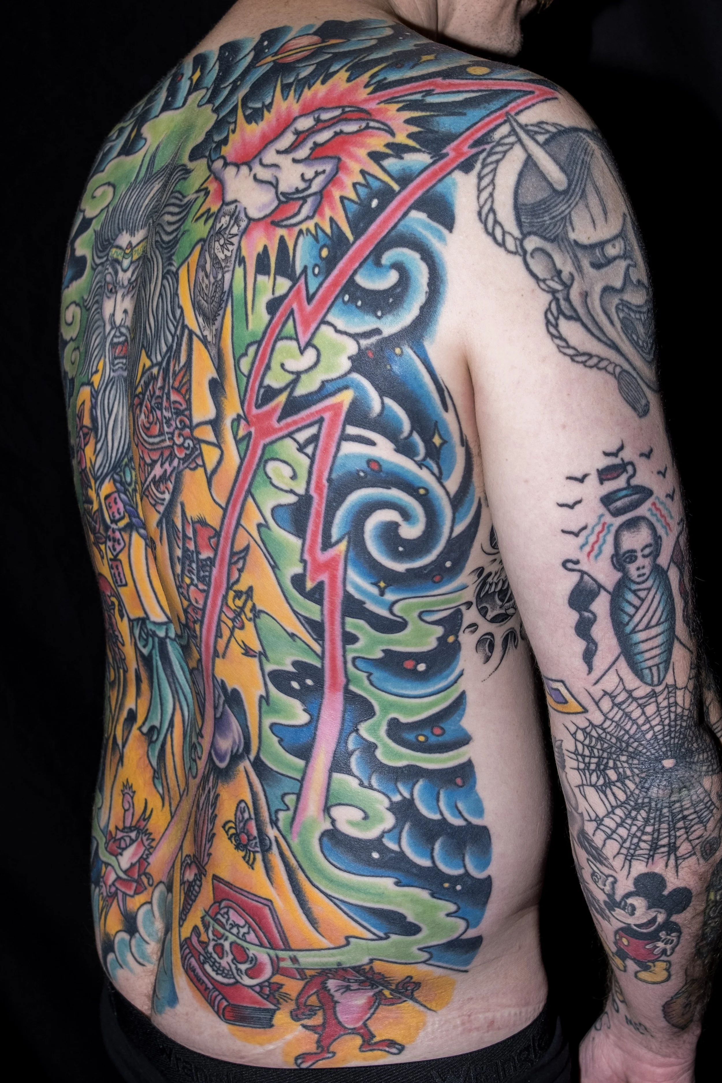 Colorful tattoo sleeve on man's chest, shoulder, and arm, featuring a lightning bolt, clouds, ocean waves, a skull, a girl holding a gun, and Japanese-style characters and imagery.