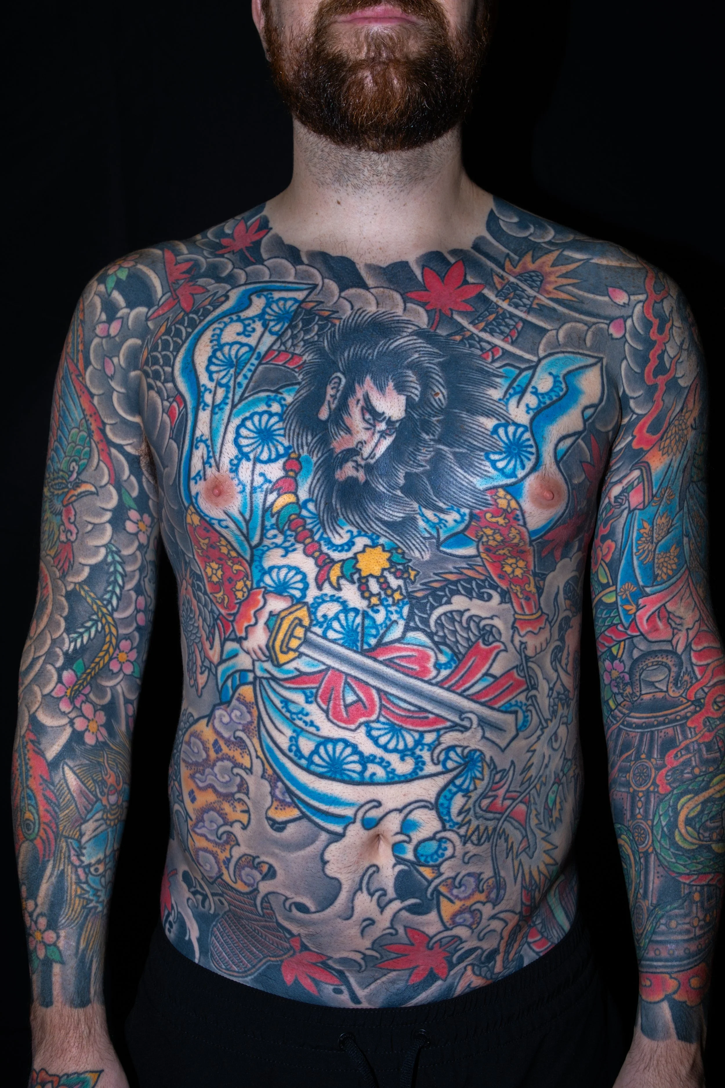 A shirtless man with a full chest and arm tattoos, featuring a prominent traditional-style dragon and a fierce lion, against a black background.