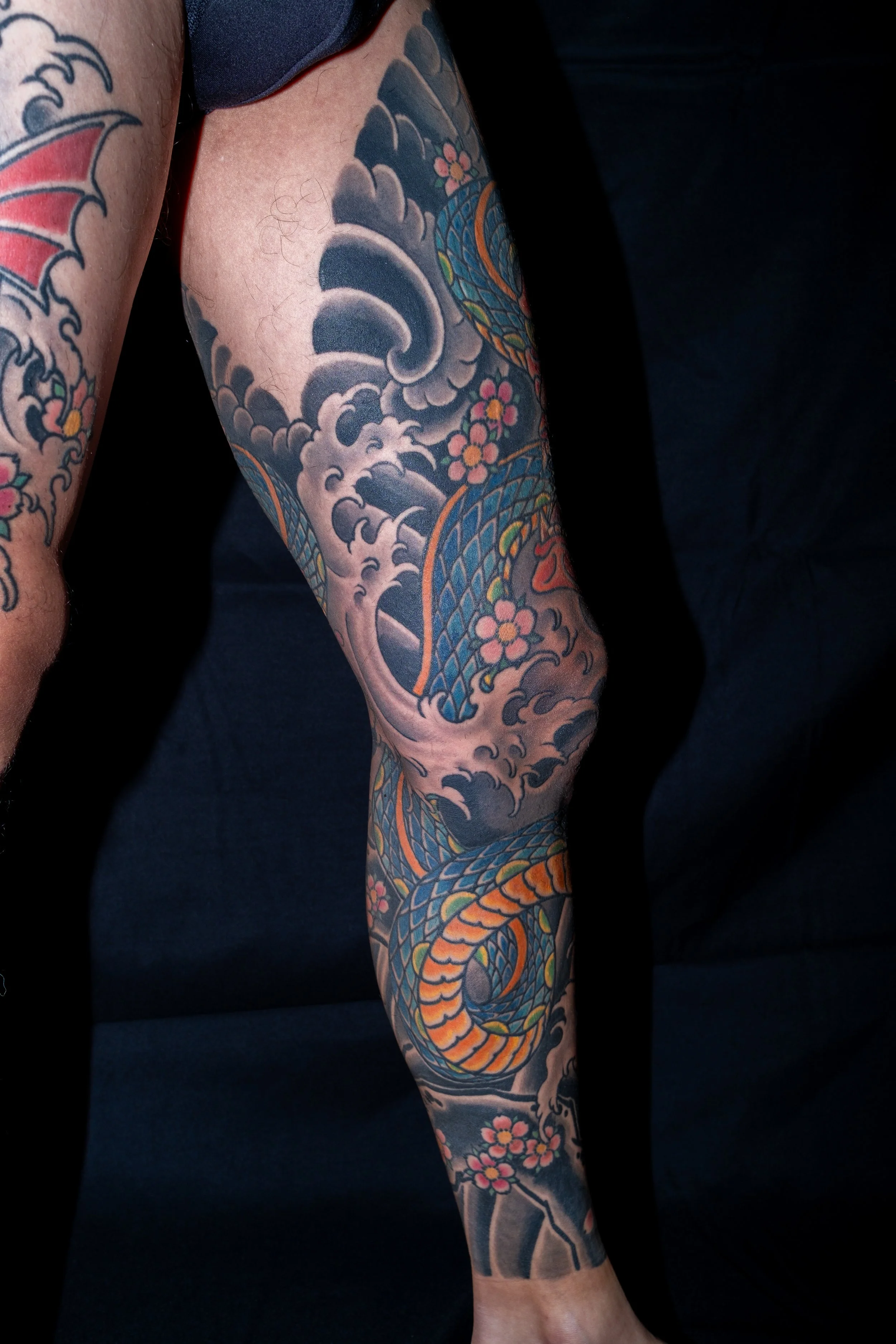 Colorful Japanese-style dragon tattoo on a person's leg, featuring the dragon's head, scales, and floral details, against a black background. Tattoo by Joe Haasch. 