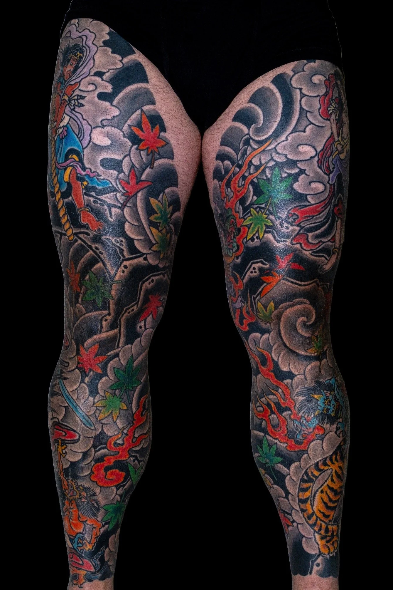 Tattooed legs with Japanese-style artwork including clouds, dragons, and maple leaves against a black background.