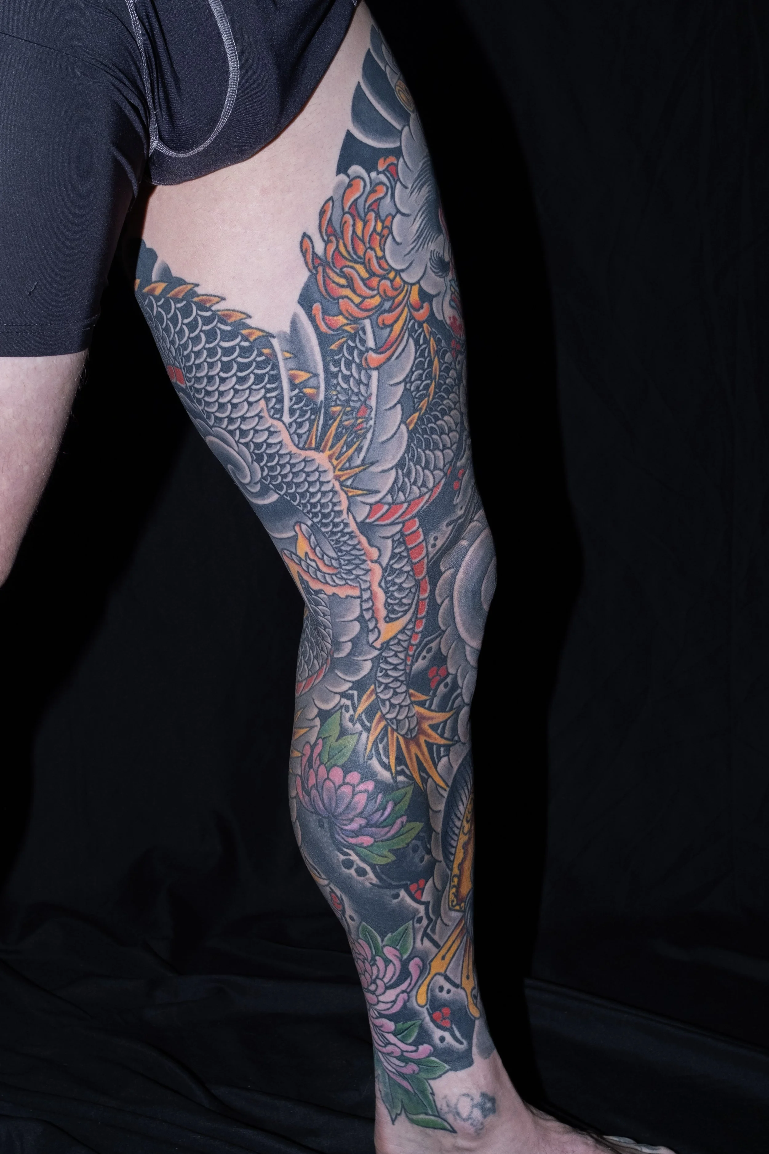 Close-up of a tattooed arm with a Japanese-style dragon and floral designs against a dark background. Tattoo by Joe Haasch. 