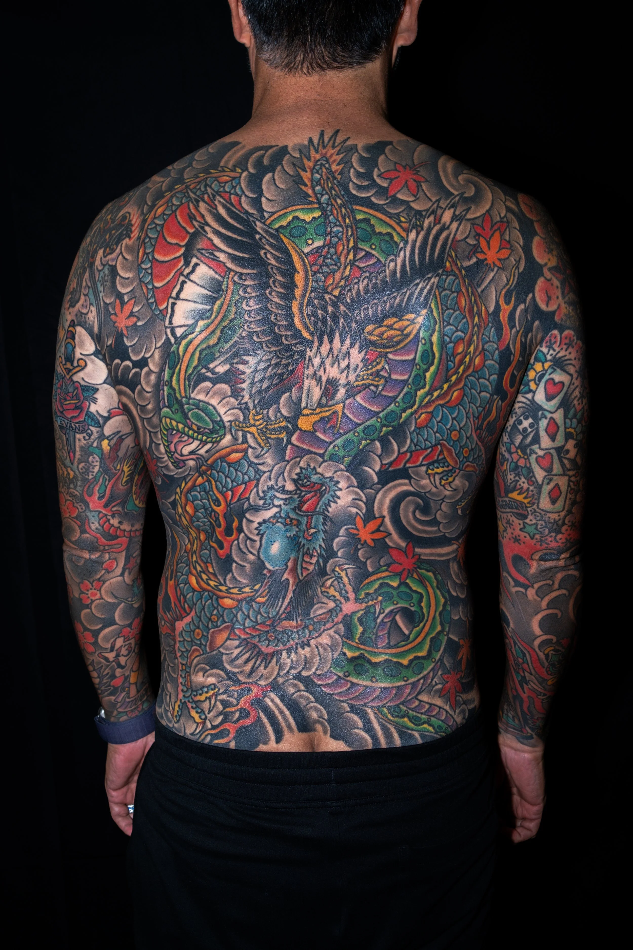 Back view of a person with detailed colorful tattoos on their back and arms, featuring a dragon, an eagle, clouds, and cherry blossoms, set against a dark background.