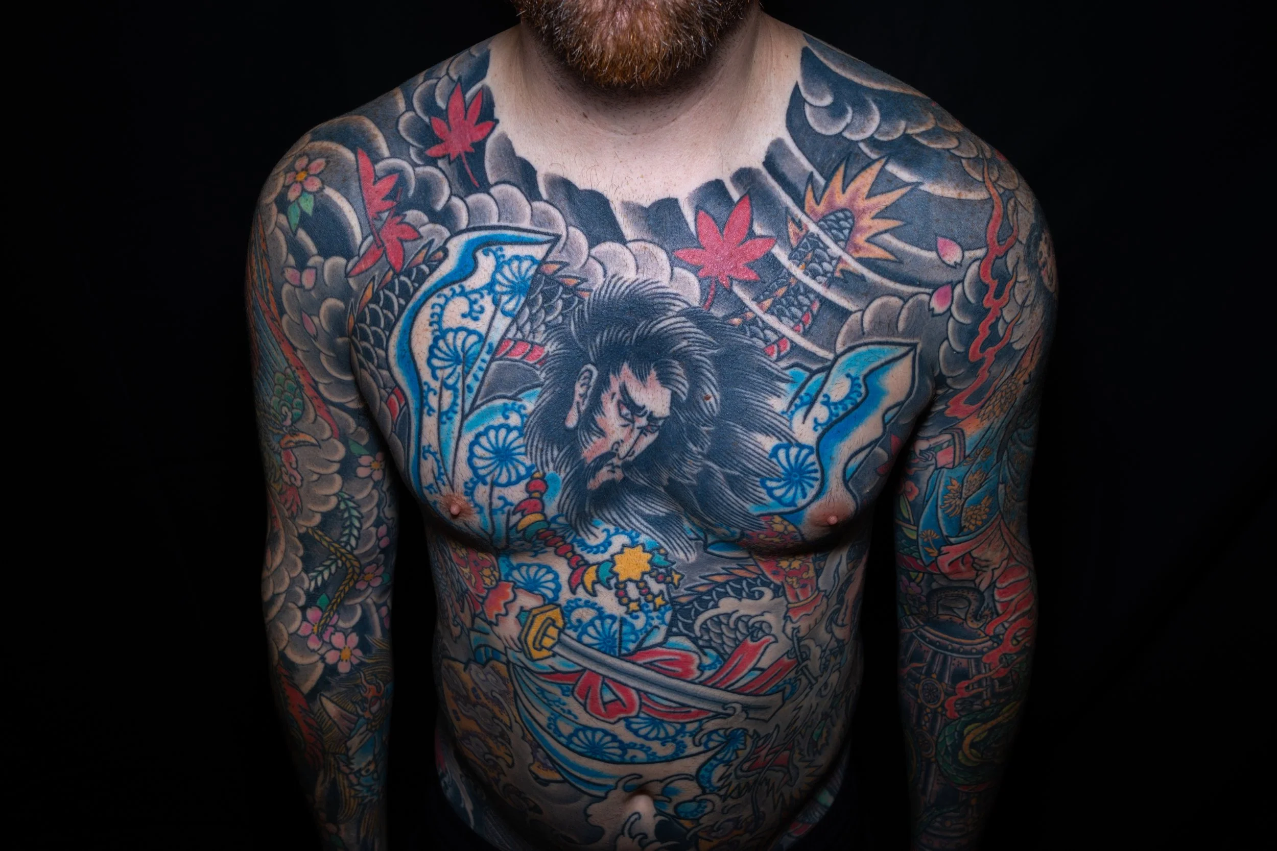 A man with extensive colorful tattoos on his chest, arms, and shoulders, standing against a black background.