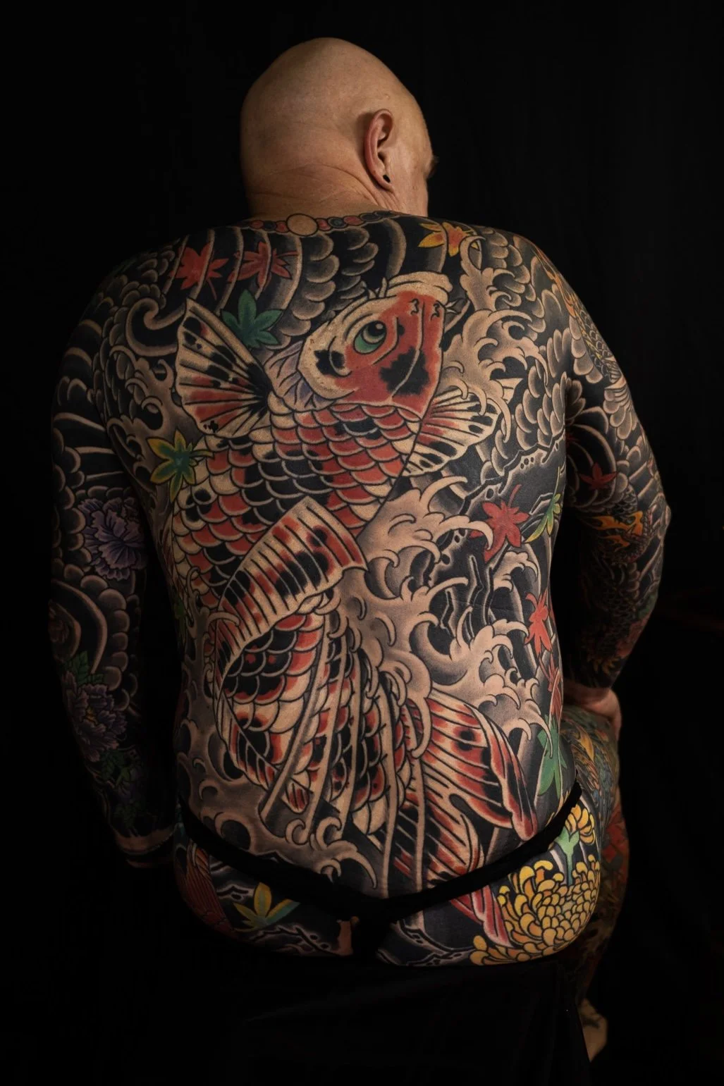 Back view of a bald man with traditional Japanese style full back tattoo of a koi fish, waves, clouds, and colorful maple leaves on dark background.
