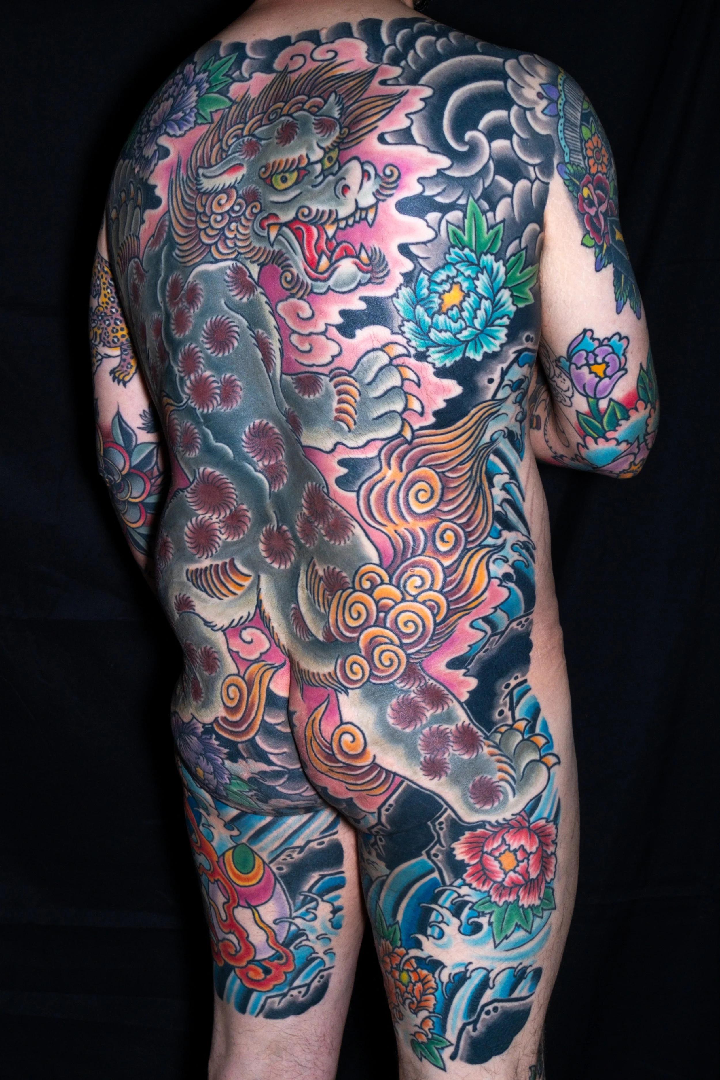 A person with intricate Japanese-style tattoos covering their back, chest, arms, and legs, featuring a large fierce dragon, colorful flowers, clouds, and waves.