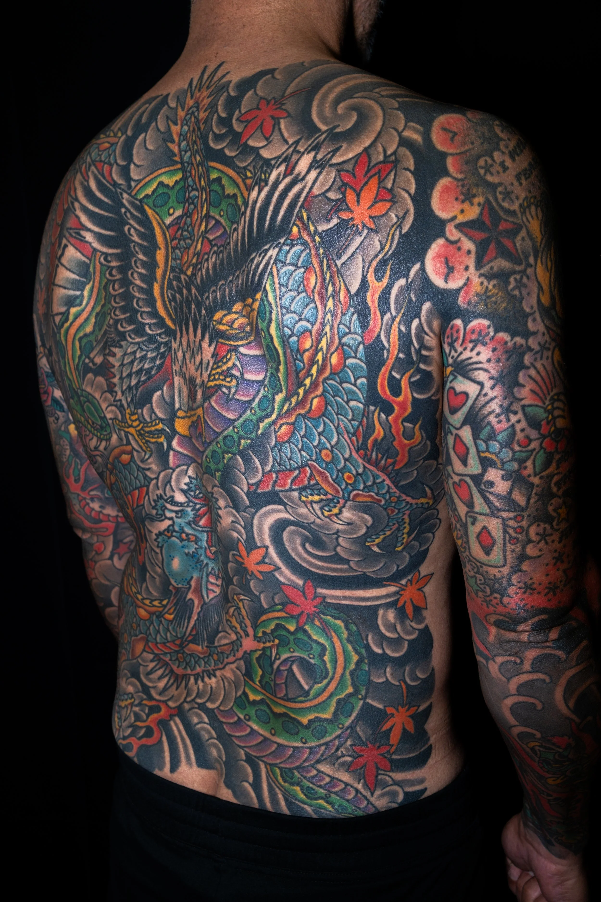 A person's torso and arm covered in colorful traditional Japanese style tattoos, including a large dragon with scales, clouds, and red maple leaves.