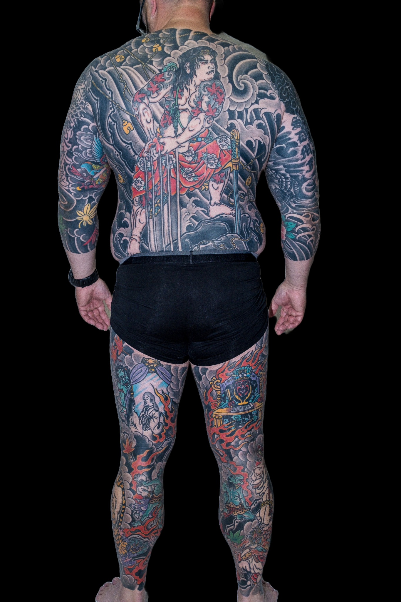 A man with extensive traditional Japanese style tattoos covering his back, arms, and legs. The tattoos depict various Japanese mythological and cultural elements, including warriors, dragons, and floral patterns.