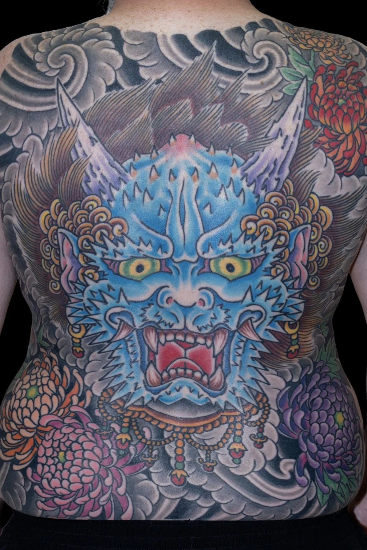 Colorful tattoo of a dragon's face on person's chest, with flames, flowers, and clouds in the background. Tattoo by Joe Haasch. 
