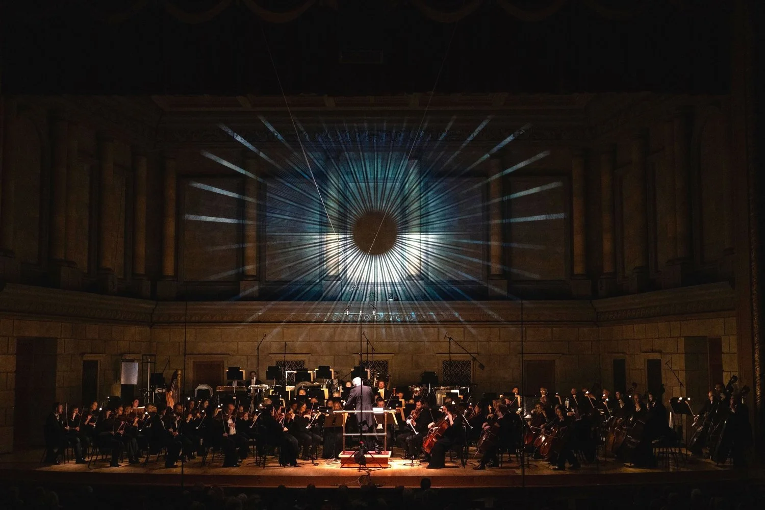 An orchestra performing on stage in a concert hall, with a large projection of a full moon and starburst pattern on the ceiling behind them.