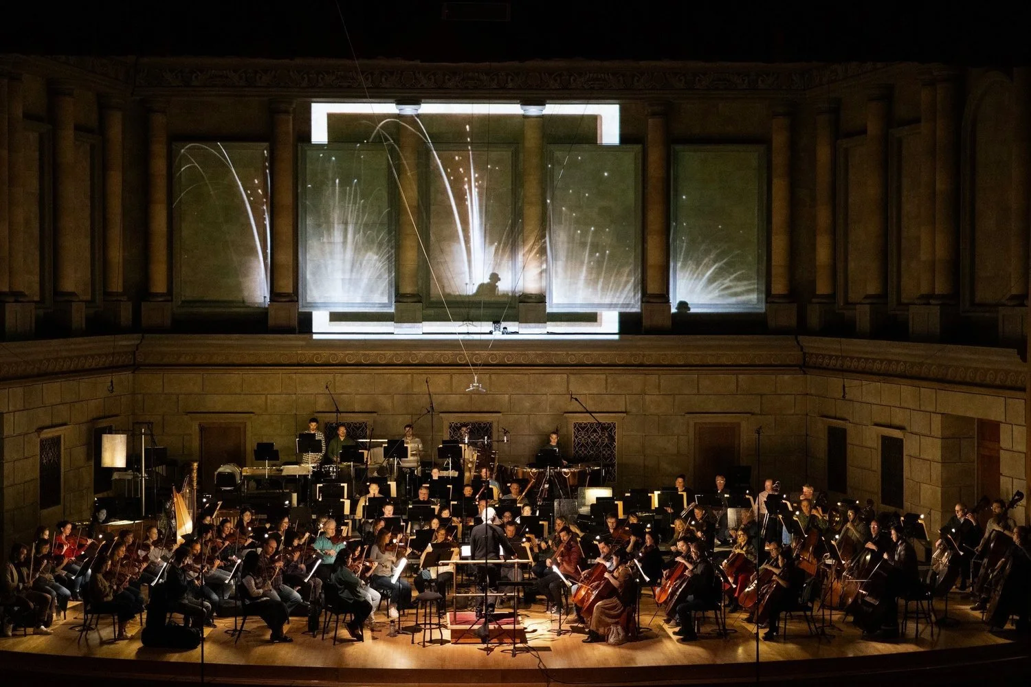 An orchestra performing on stage in a concert hall, with a large screen behind displaying light effects, and musicians with their instruments seated in front of a conductor.