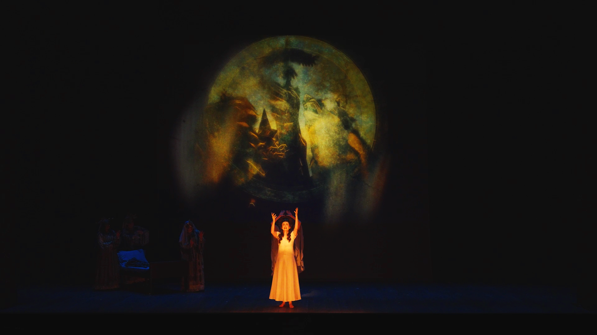 Performers on stage during a theatrical production, with a woman in a beige dress raising her arms, and a large circular backdrop projecting an abstract image.
