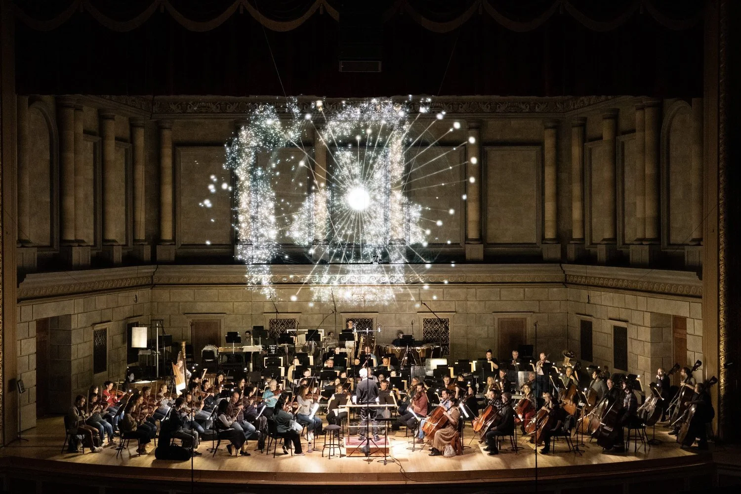 An orchestra performing on stage with a digital firework display in the background, inside a concert hall with a wooden stage and ornate wall details.