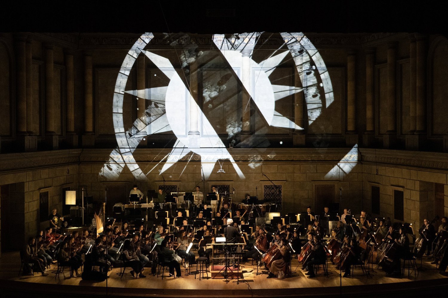An orchestra performing on stage in a concert hall with large geometric light projections on the wall behind them.