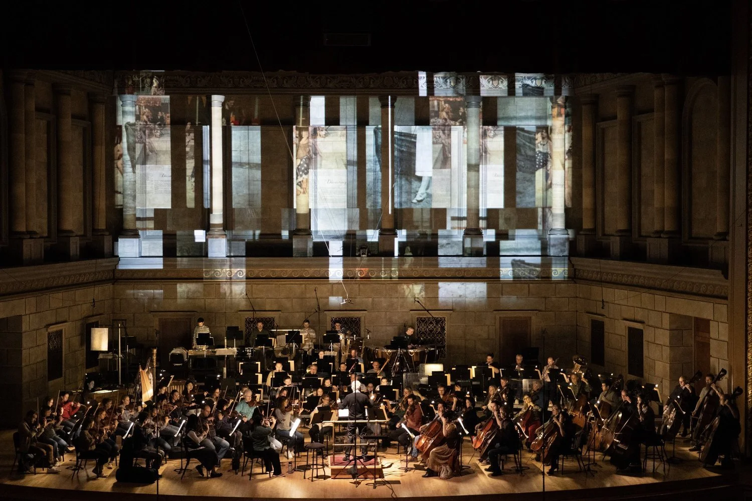 Orchestra performing on stage in a concert hall with projected images on the backdrop.