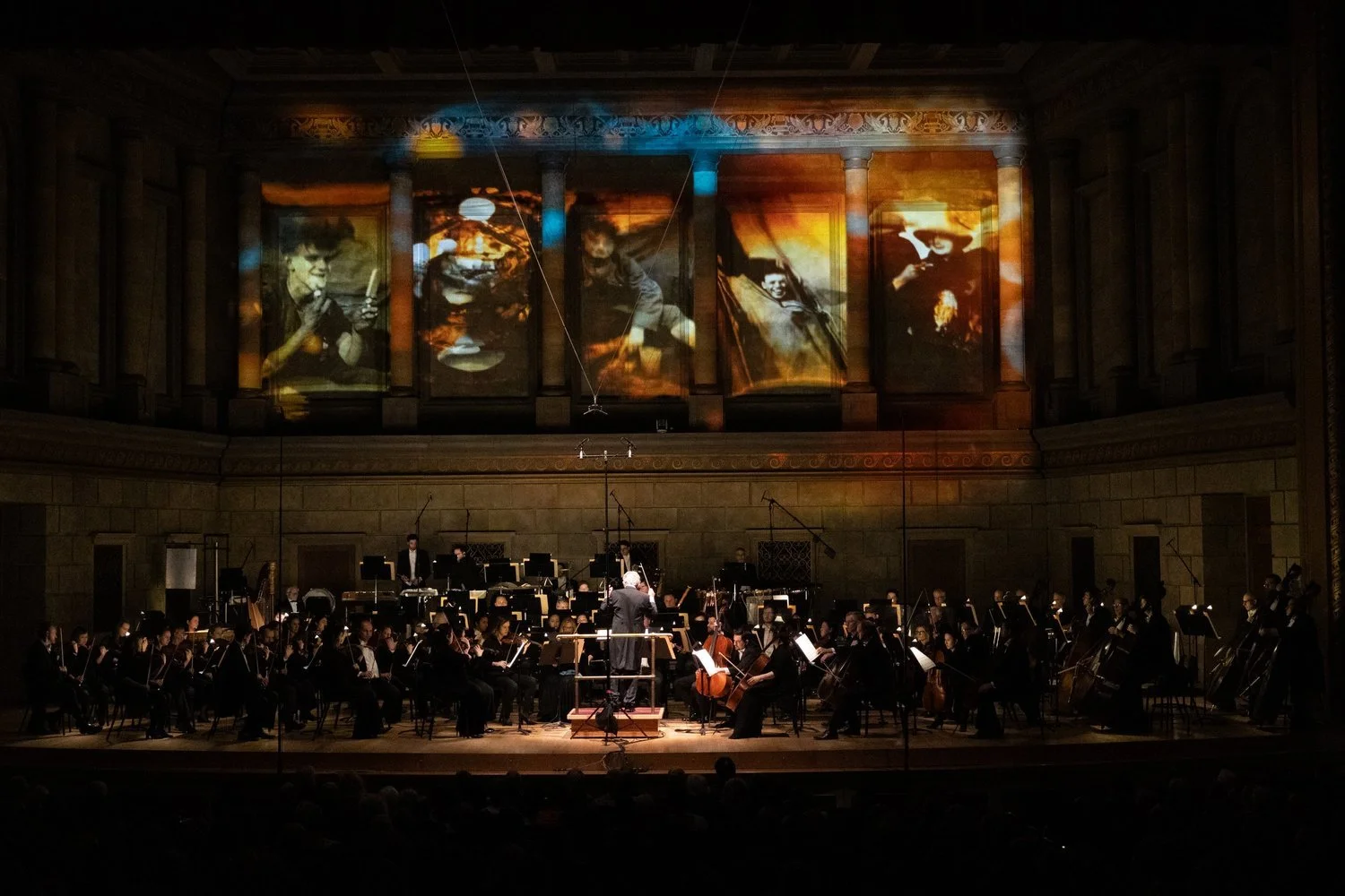 An orchestra performing on stage in a concert hall, with four large abstract paintings projected above them.