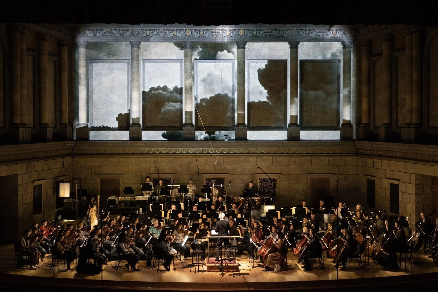 An orchestra performing on stage with a backdrop of sky views and clouds.