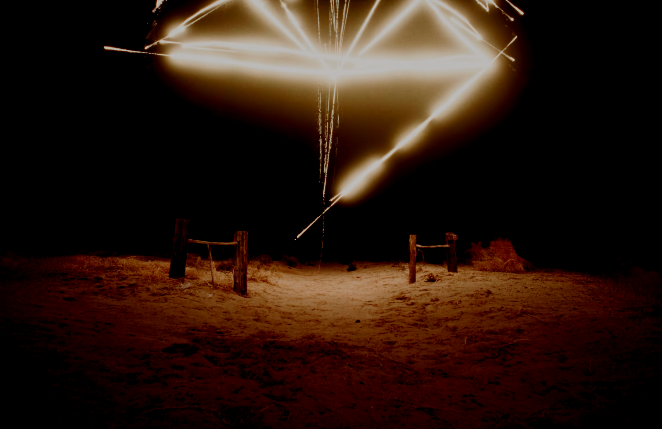 Light Drawing illiuminating the night sky over a sandy outdoor area with wooden posts.