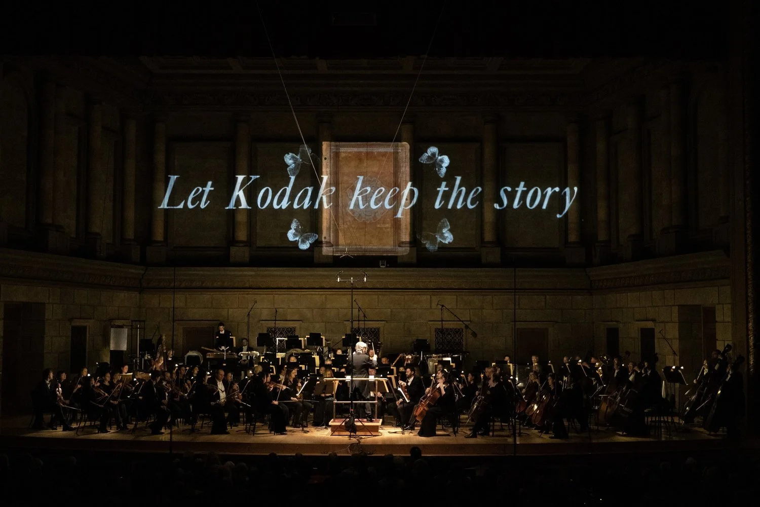 An orchestra performing on stage in a concert hall with a projection behind them that reads 'Let Kodak keep the story' and has butterfly graphics.