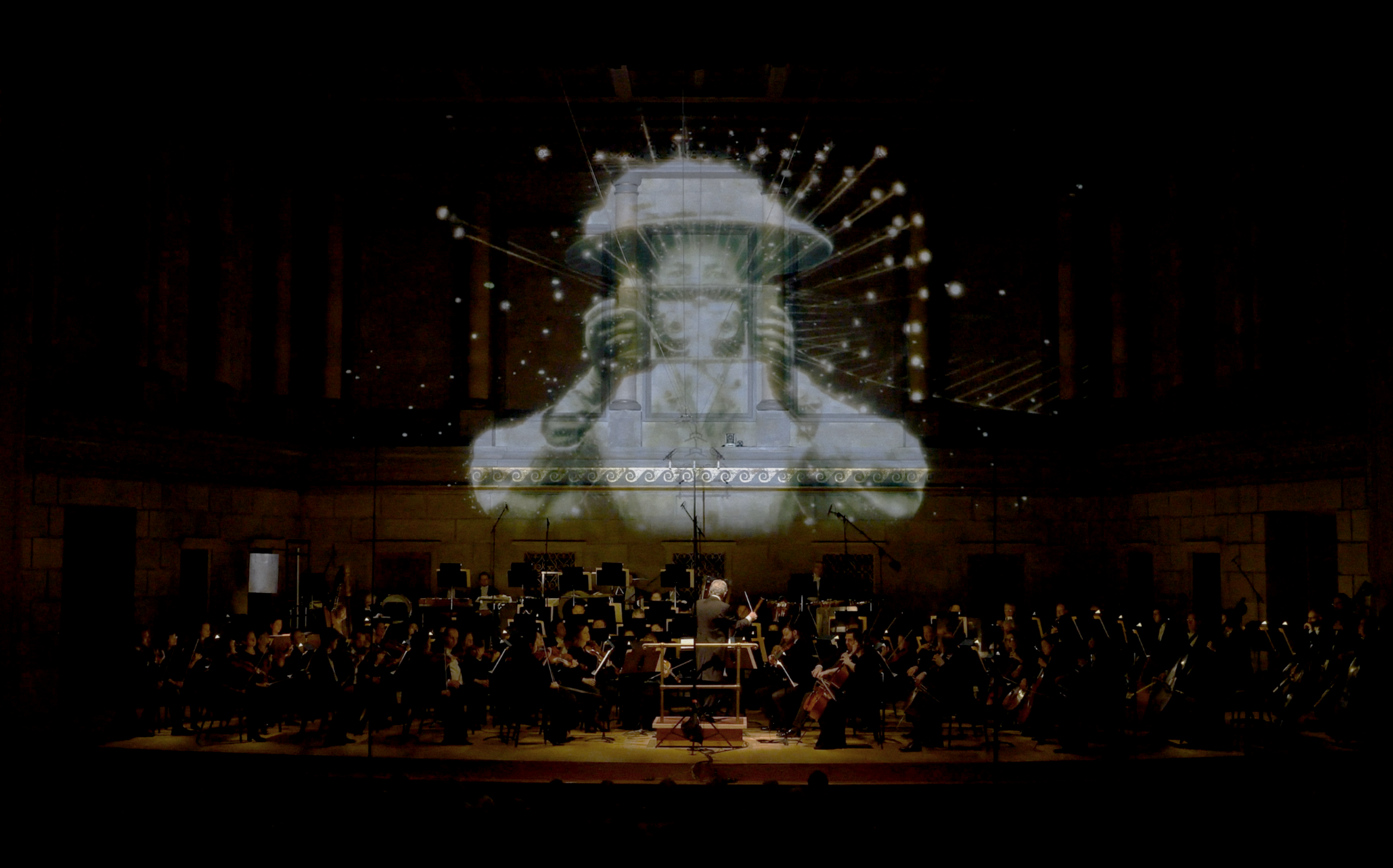 Orchestra performing in a concert hall with projected visuals of a person holding a top hat with the Earth in the background.