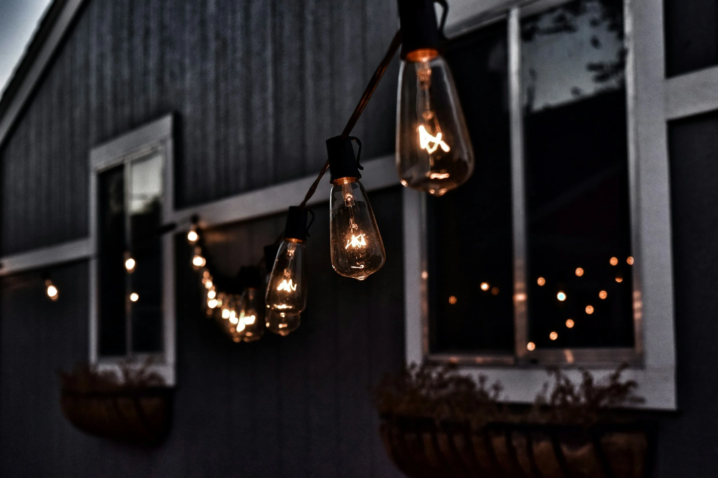 String of hanging Edison-style light bulbs illuminated at night outside a house with large windows and a porch.
