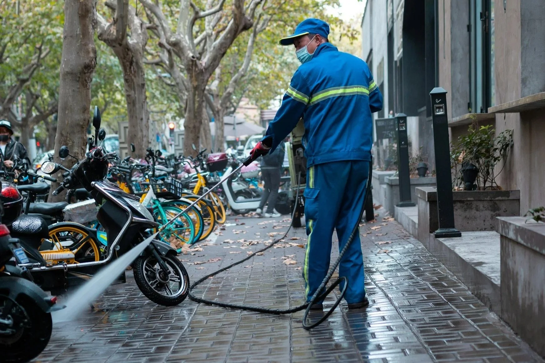 A worker in a blue uniform and face mask using a pressure washer to clean a sidewalk in an urban area with parked bicycles and trees.