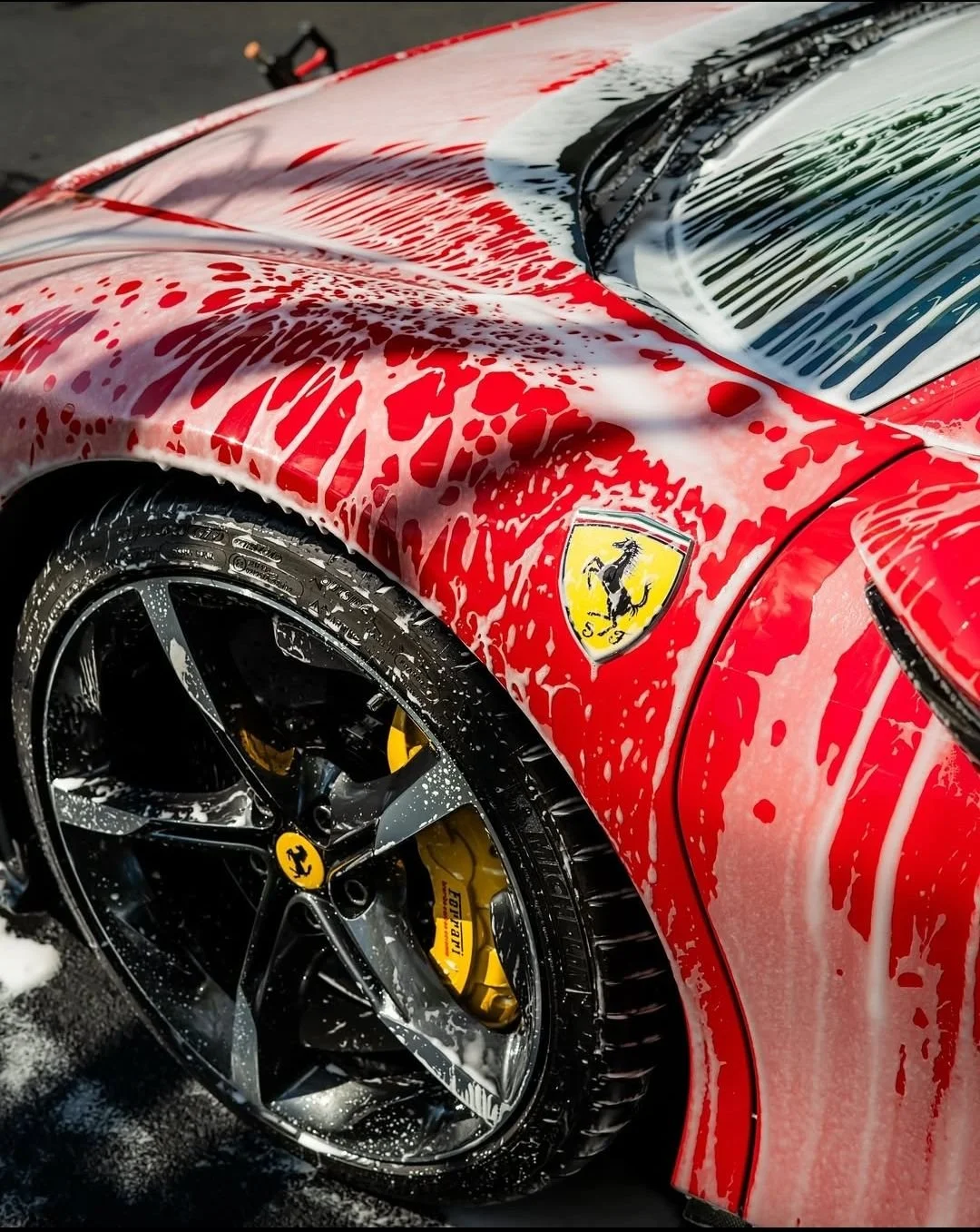 Close-up of a red Ferrari sports car, covered in soap suds during washing. The Ferrari logo is visible on the front side panel, and the wheel features a yellow brake caliper with the Ferrari name.
