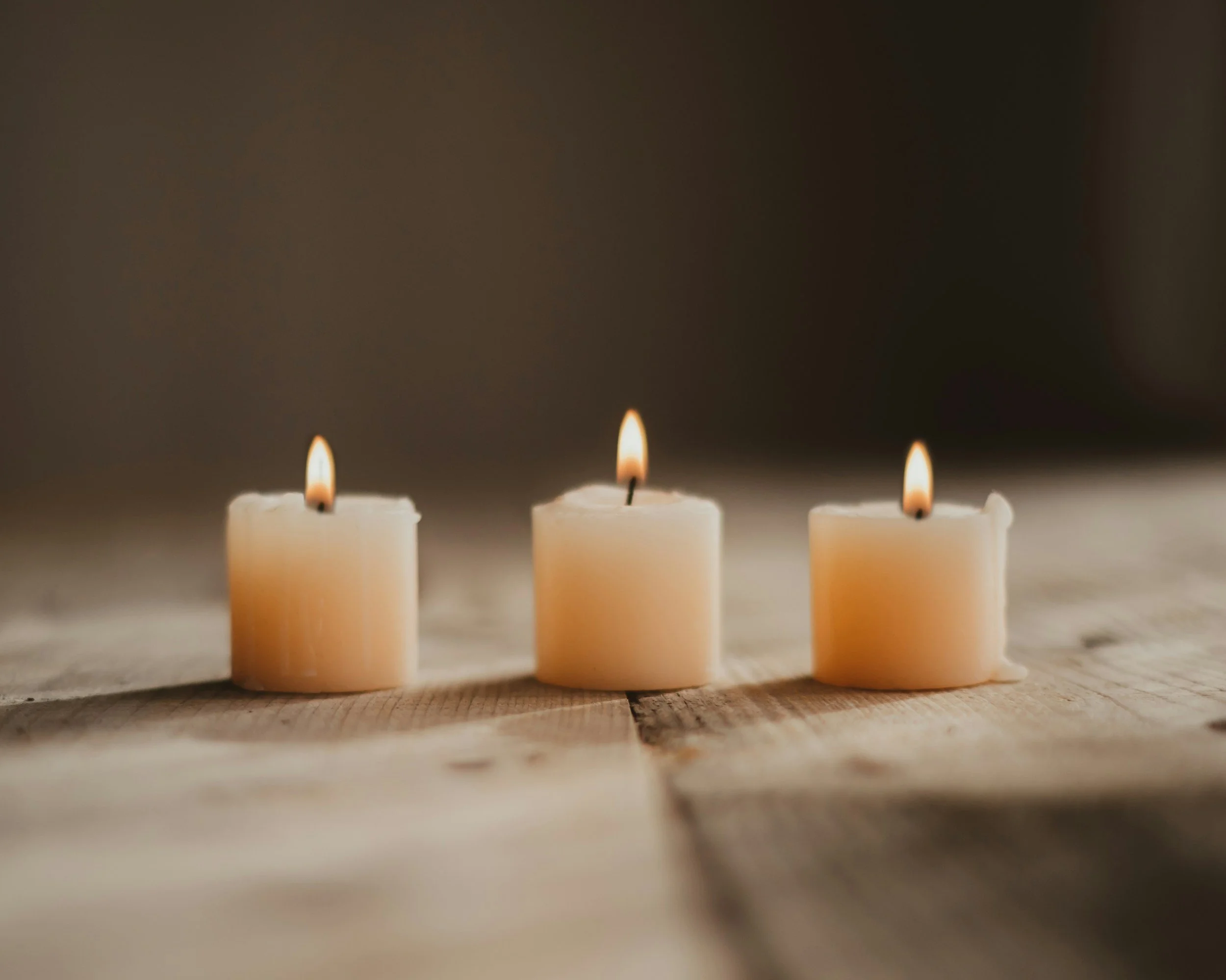 Three white candles burning on a wooden surface with a blurred dark background.