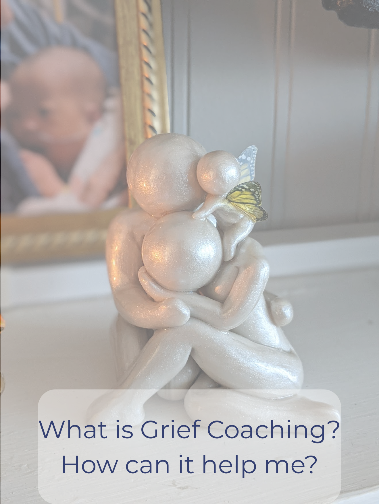 A decorative figurine of a person sitting with legs crossed, holding a cluster of shiny, pearl-like spheres. The figurine has wings and a butterfly resting on one of the spheres. In the background, there is a blurred framed photograph of a child or young person with the words "What is Grief Coaching? How can it help me?" written at the bottom.