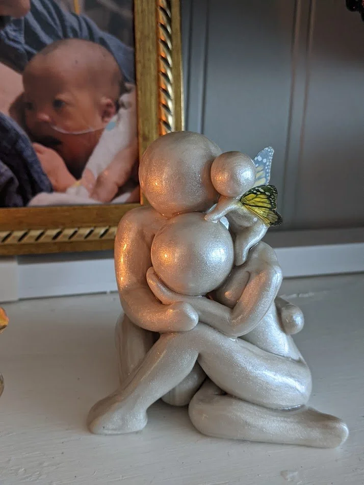 Silver-colored sculpture of two abstract human figures embracing with a small butterfly baby figure perched on them, signifying child loss, in front of a family photo of a baby with a nasal cannula.