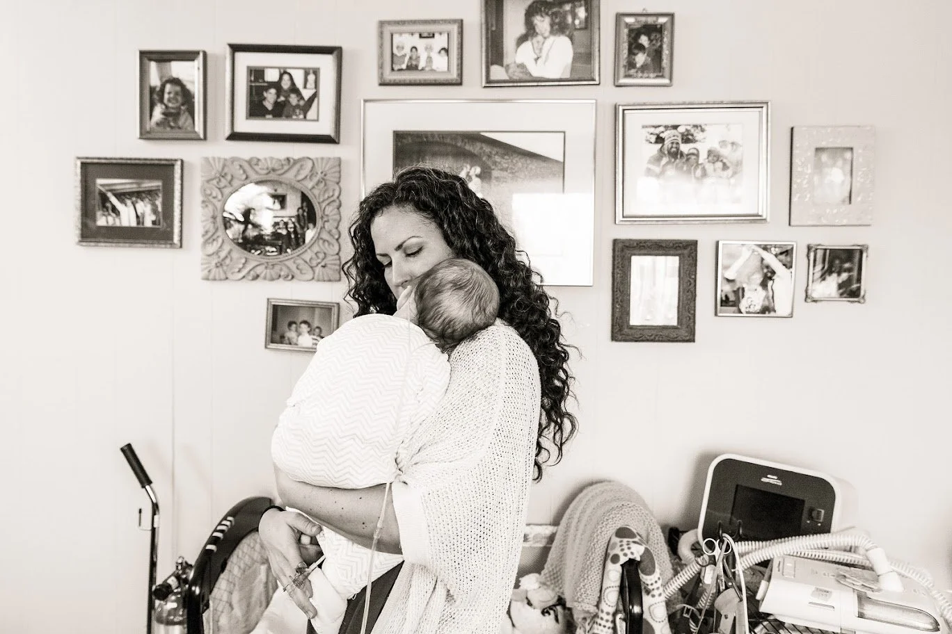 A woman with curly hair holding a baby in a hospital room with framed photos on the wall.