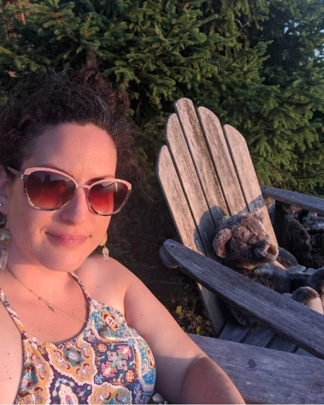 Emily, grief coach, wearing sunglasses and a patterned tank top sitting at the beach at sunset next to a stuffed bear lying on a wooden Adirondack chair, with green trees in the background.