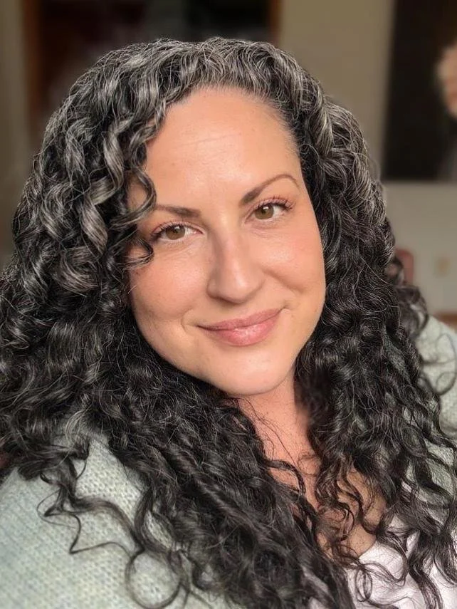 Emily's portrait, smiling gently toward the camera with dark curly hair and brown eyes, and a soft blue sweater.
