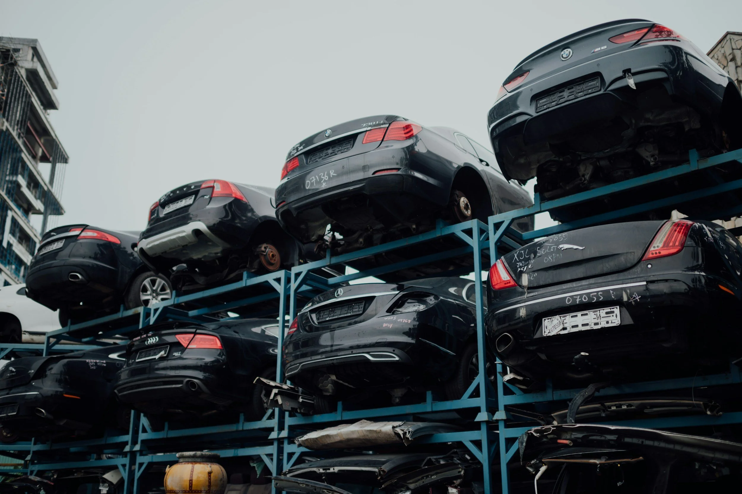 Several black cars stacked on a blue metal rack at a salvage yard, some with visible damage and missing parts, under an overcast sky.