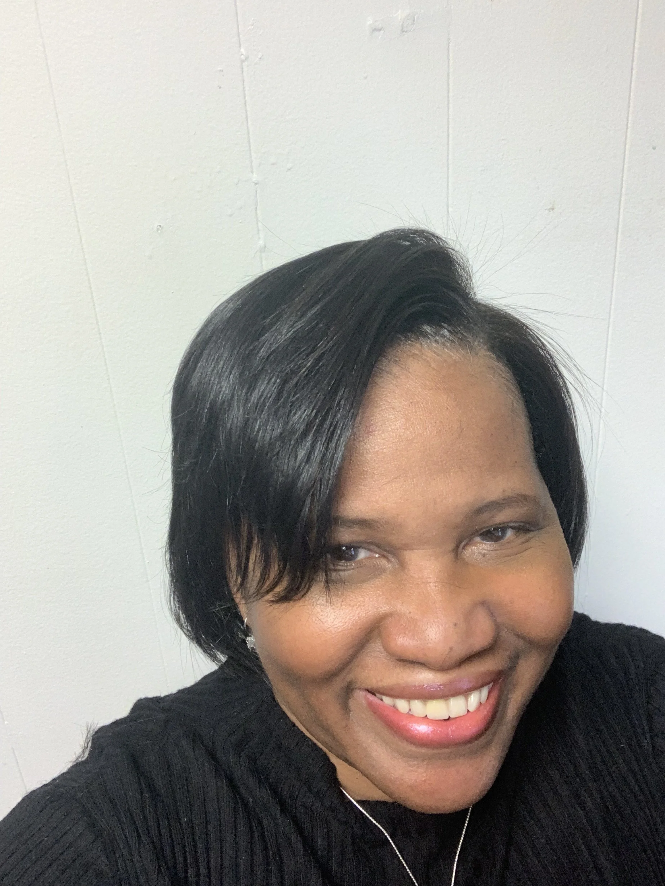 A smiling woman with short black hair, wearing a black top and earrings, against a white wall.
