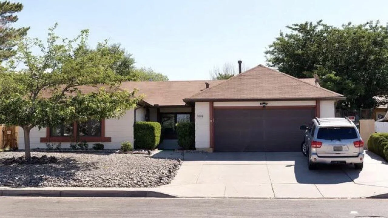 Single-story house with tan roof, white walls, brown trim, two front windows, a closed garage door, and a silver SUV parked on the driveway. There are trees, bushes, and rocks in the front yard.