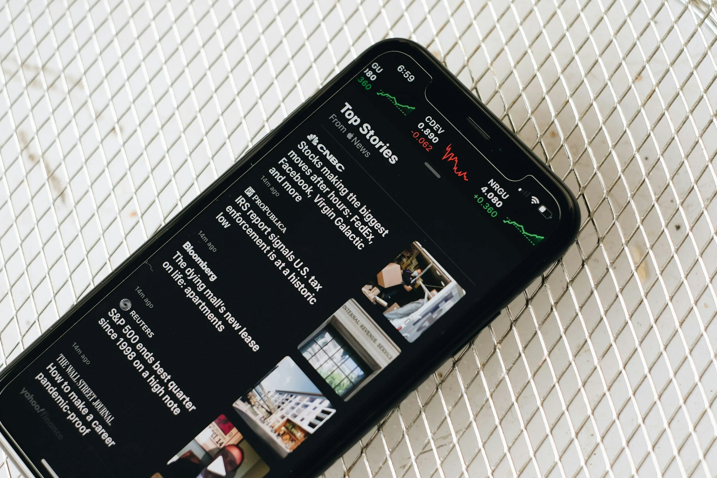 Close-up of a smartphone displaying news headlines on a black background, placed on a metal mesh surface.