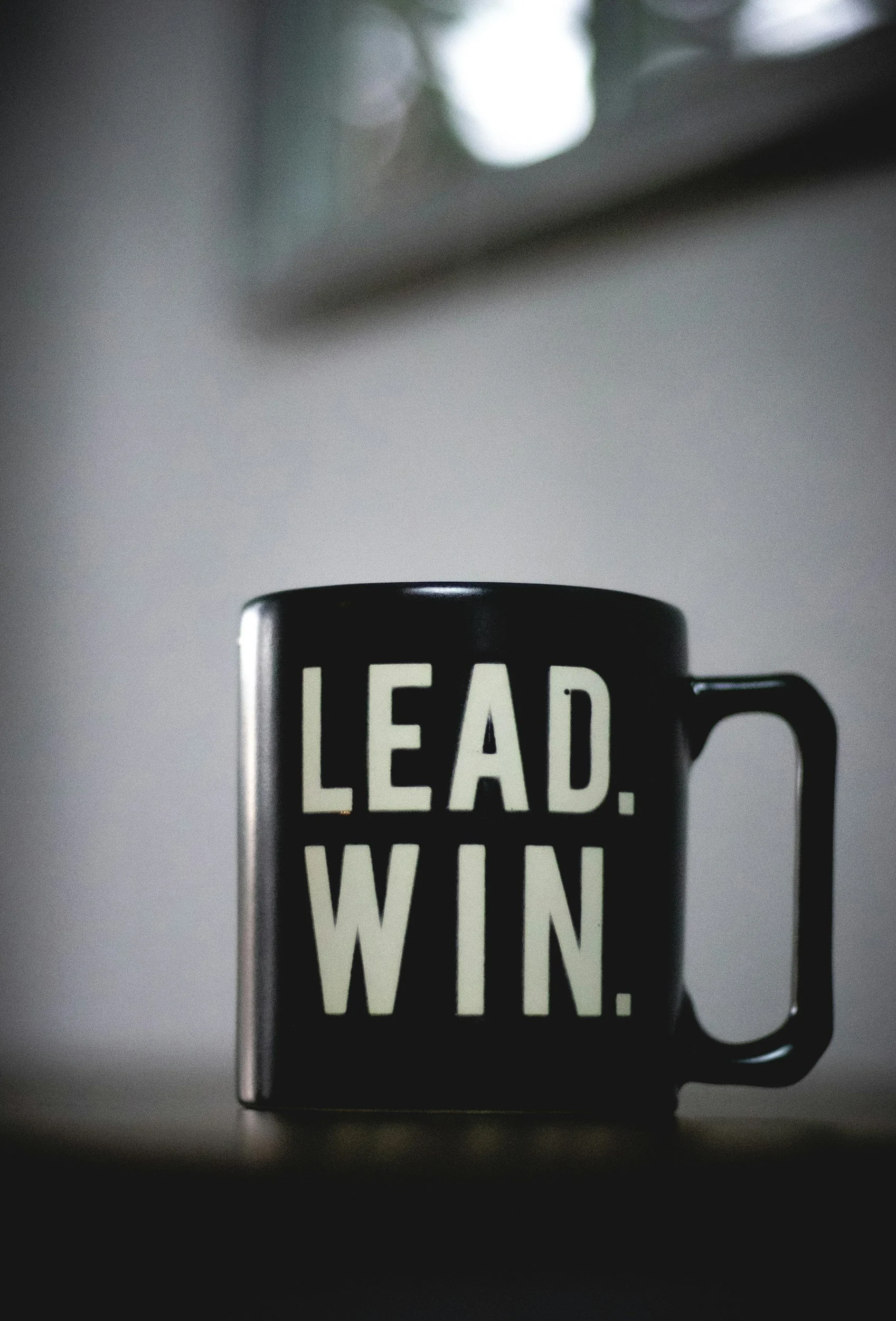 A black coffee mug with bold white text reading 'LEAD. WIN. WIN.' placed on a dark surface with a blurred background.
