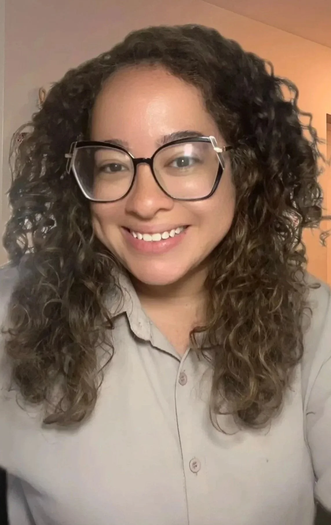 A smiling woman with curly brown hair, wearing large black glasses and a light beige button-up shirt.