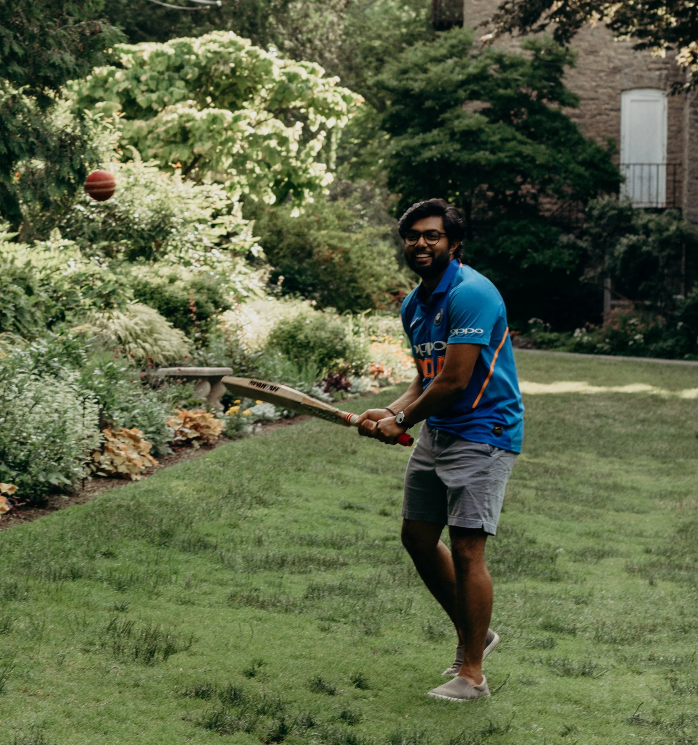 Young man with glasses and beard playing cricket outdoors in a garden with lush greenery and a brick house in the background.