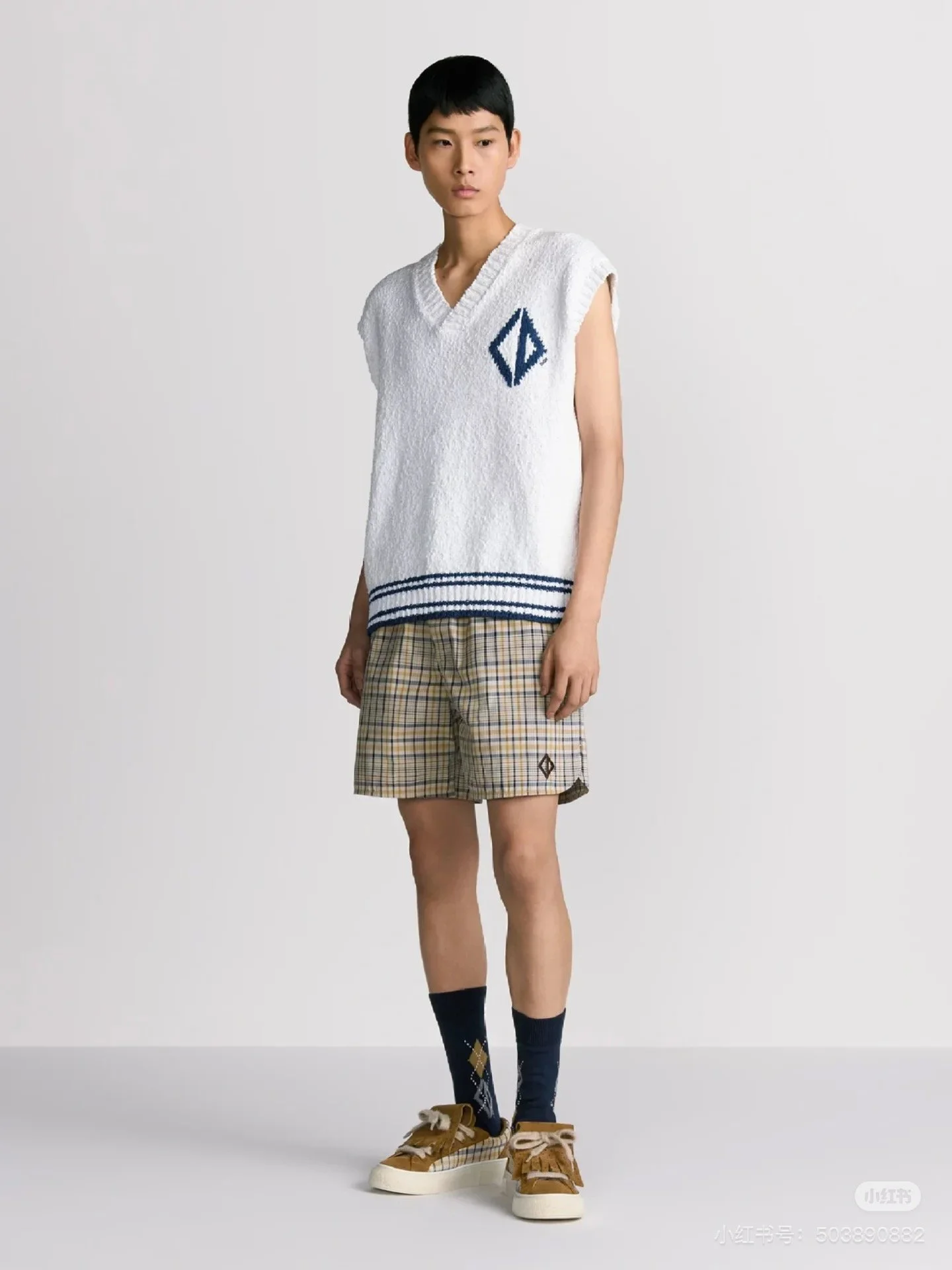 A person wearing a white sleeveless sweater with a blue geometric design on the chest, plaid shorts, dark navy socks with a diamond pattern, and yellow sneakers, standing against a plain gray background.