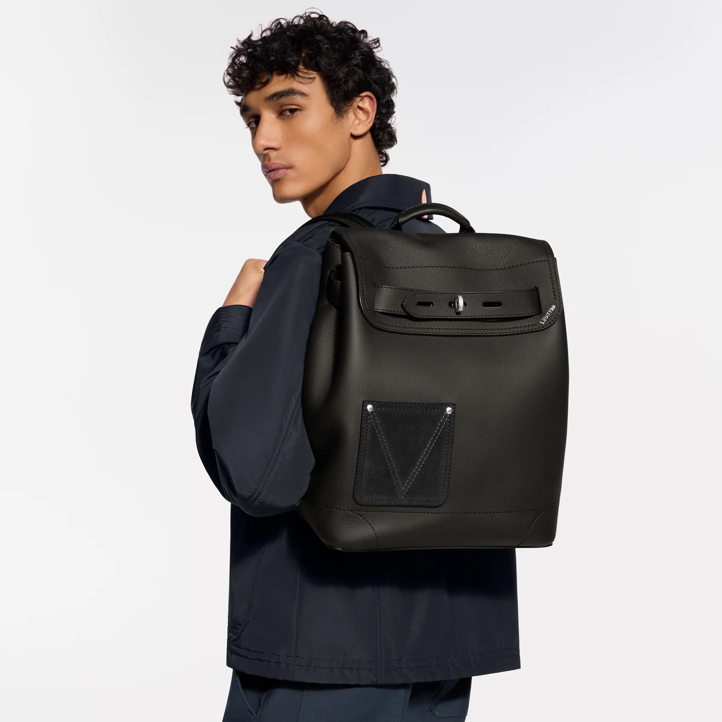 A young man with curly dark hair, wearing dark clothing and carrying a black leather backpack, looking over his shoulder against a plain white background.