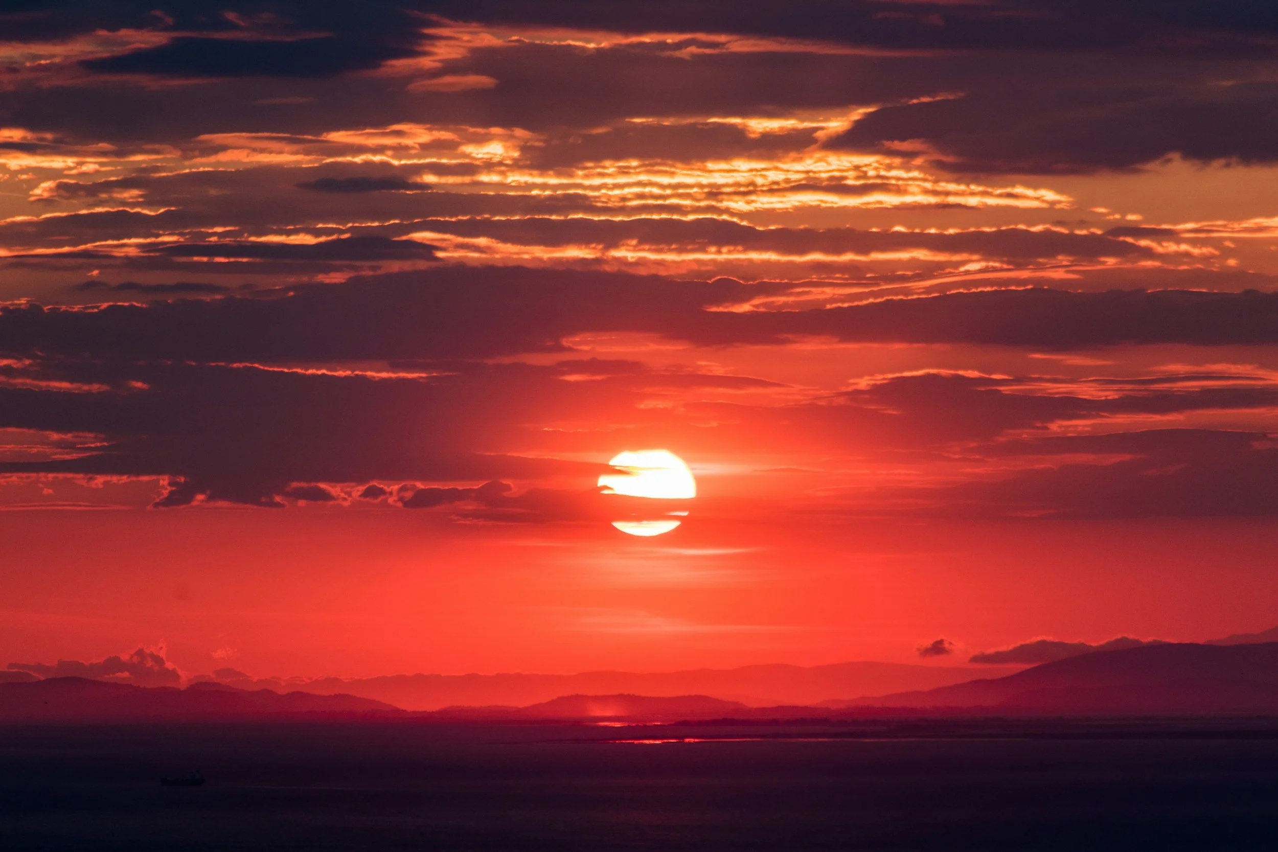 A vibrant sunset over the ocean, with the sun partially below the horizon, casting orange, pink, and purple hues across the sky and reflecting on the water below.