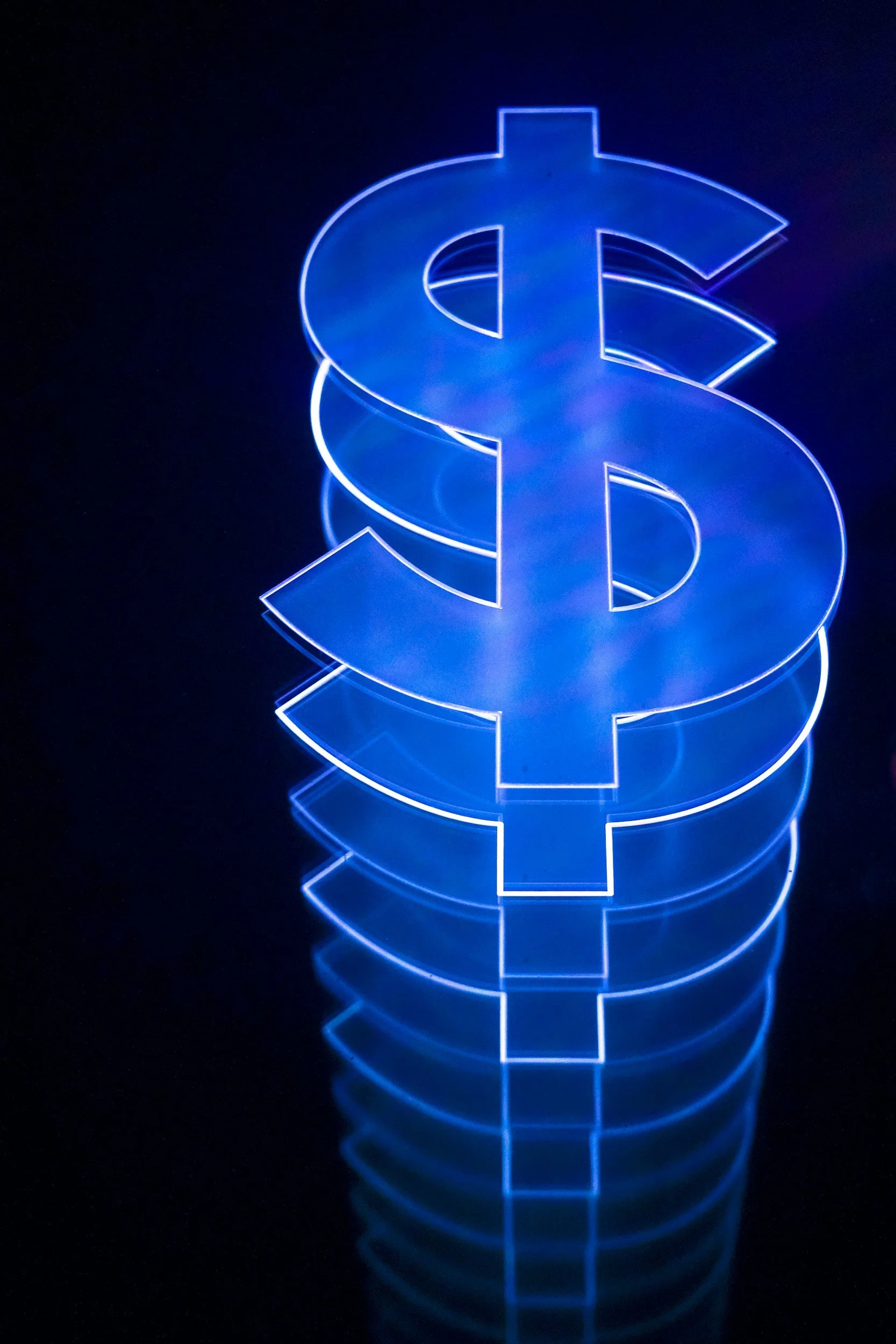 A glowing blue neon dollar sign ($) illuminated against a dark background, with its reflection visible on the shiny surface below.