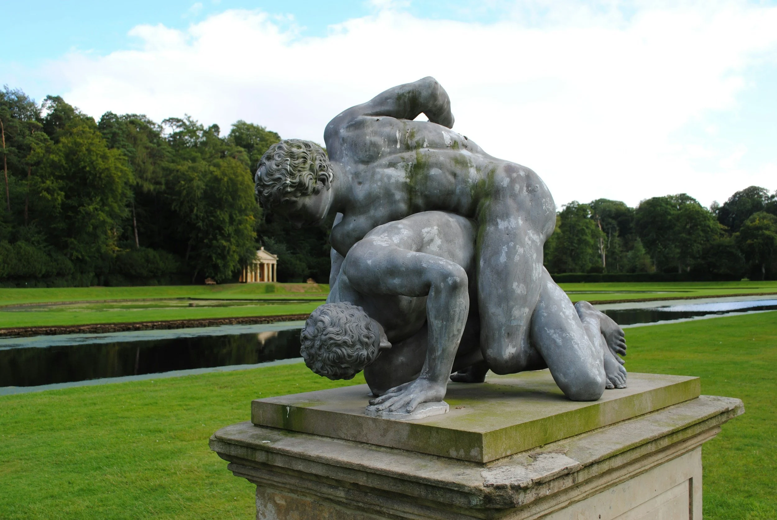 Marble sculpture of two muscular men fighting, set outdoors with trees and a building in the background.