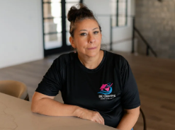 Woman with dark hair in a bun, wearing a black T-shirt with a logo, sitting at a table in a modern indoor setting.