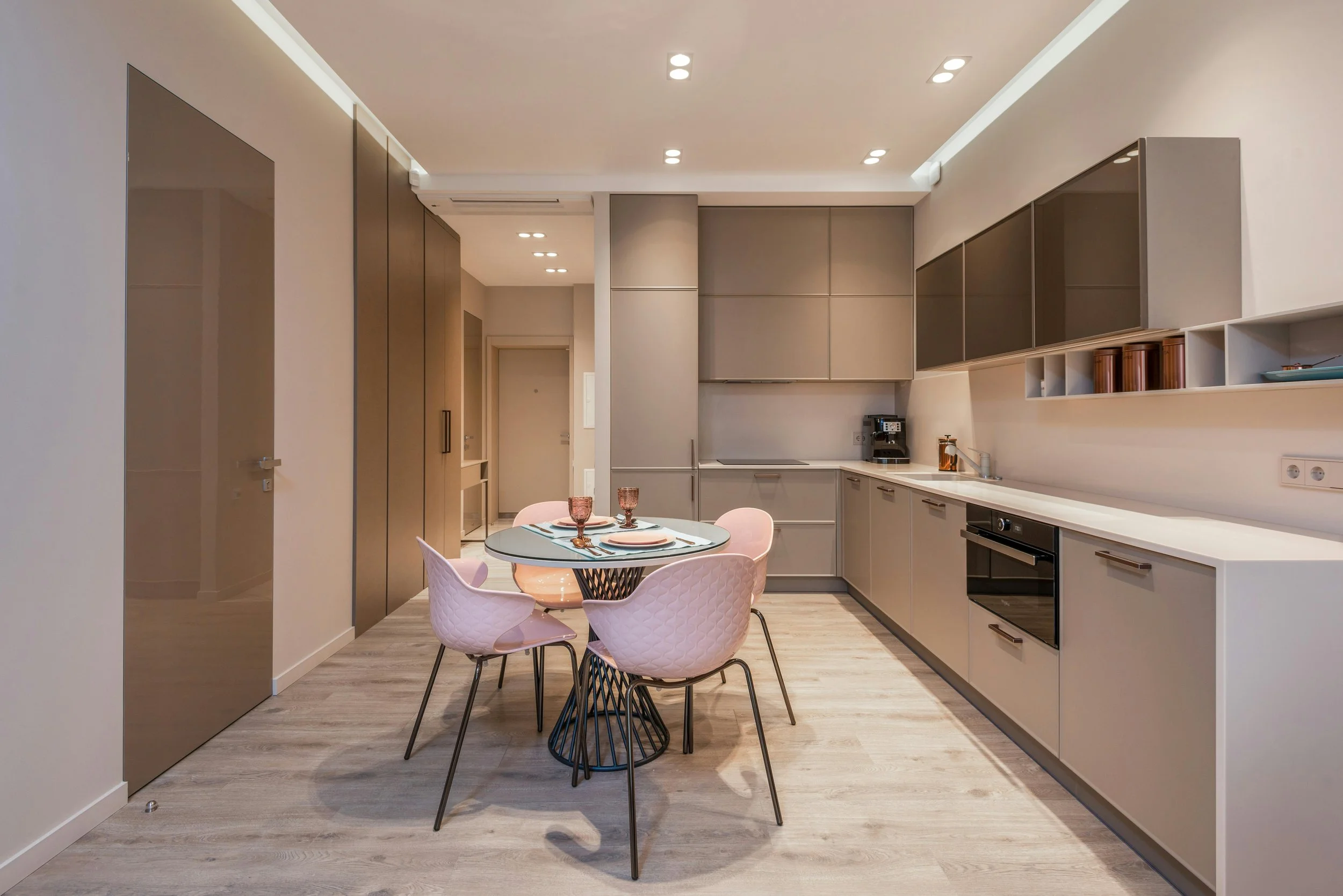 Modern kitchen with beige cabinetry, built-in oven, and a small round dining table with pink quilted chairs set for four people.